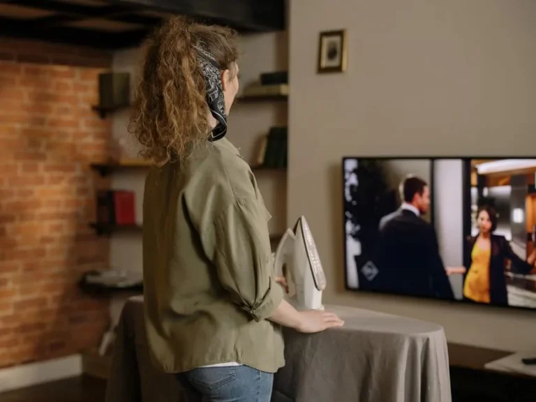 A woman ironing while watching TV in a cozy brick-walled living room.